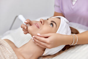 A woman lying on a treatment bed receives a facial procedure with a handheld device from a person wearing a light purple uniform.