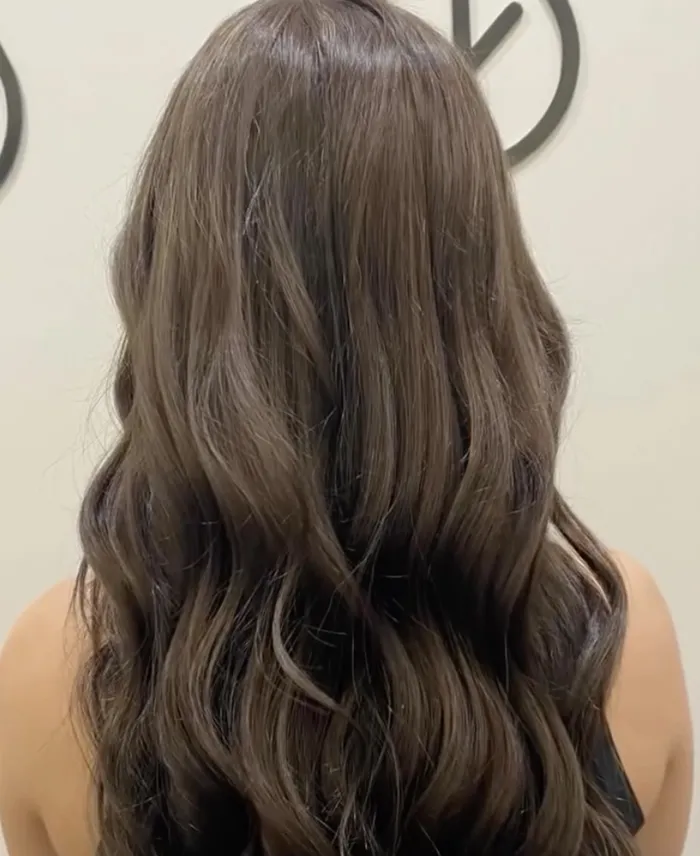 A person with long, wavy brown hair is shown from the back, standing indoors against a light-colored wall with partial circular designs. Copperfalls Aveda Spa & Salon, Castle Rock, CO