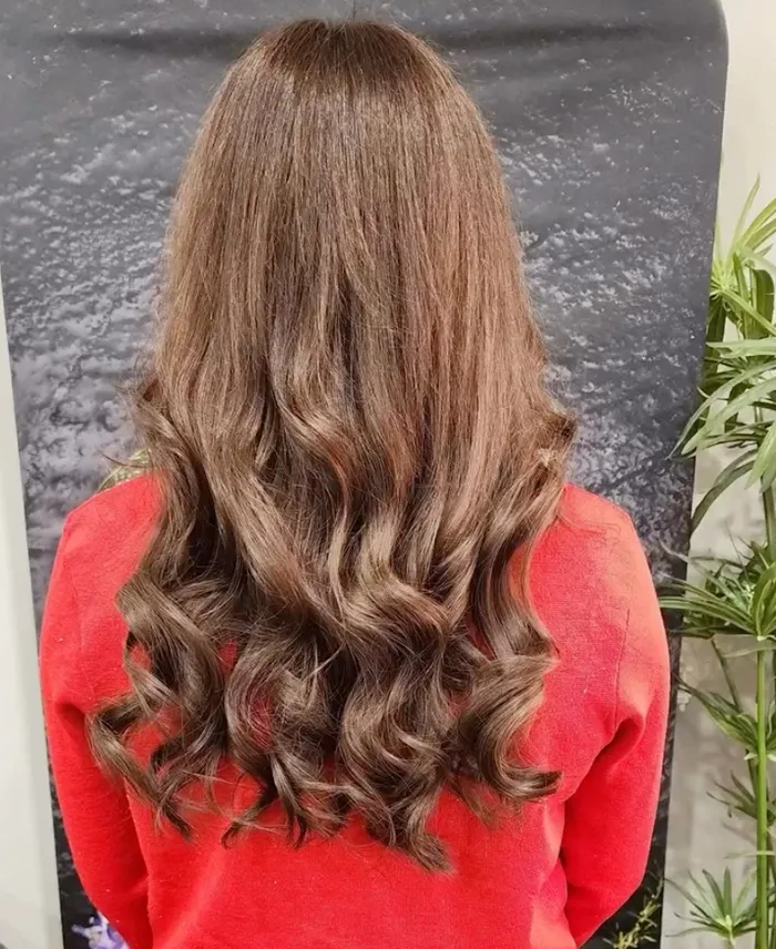 Person with long, wavy brown hair viewed from behind, wearing a red top, standing in front of a dark background with green plants on the side. Copperfalls Aveda Spa & Salon, Castle Rock, CO