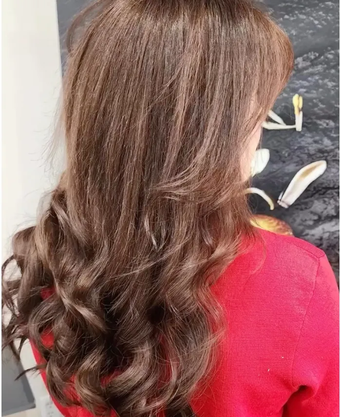Woman with long, wavy brown hair wearing a red top, photographed from the back against a dark background with abstract art. Copperfalls Aveda Spa & Salon, Castle Rock, CO