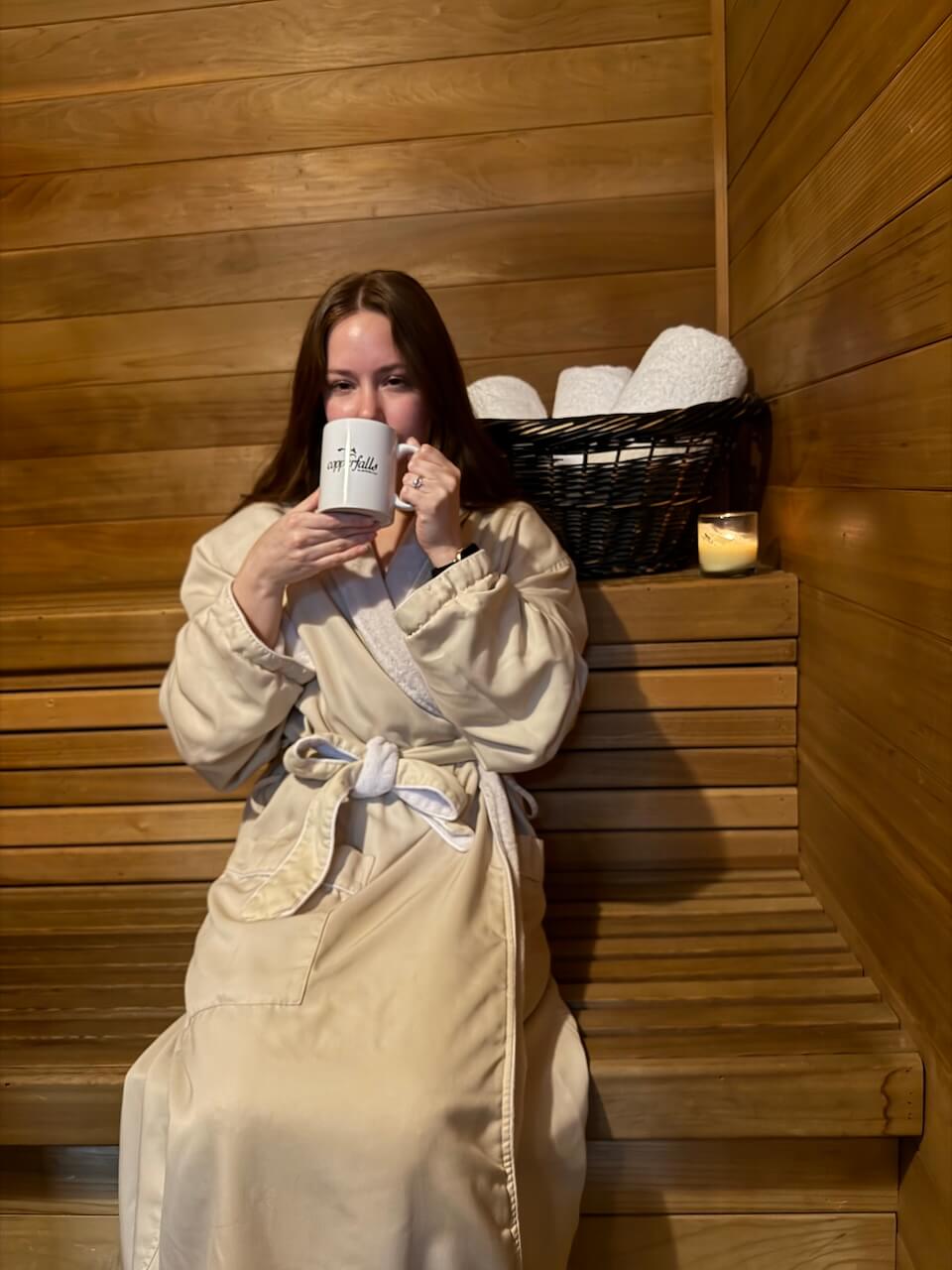 Woman in a beige robe sits on a wooden sauna bench, holding a mug. A basket of towels and a lit candle are on the bench beside her. Copperfalls Aveda Spa & Salon, Castle Rock, CO