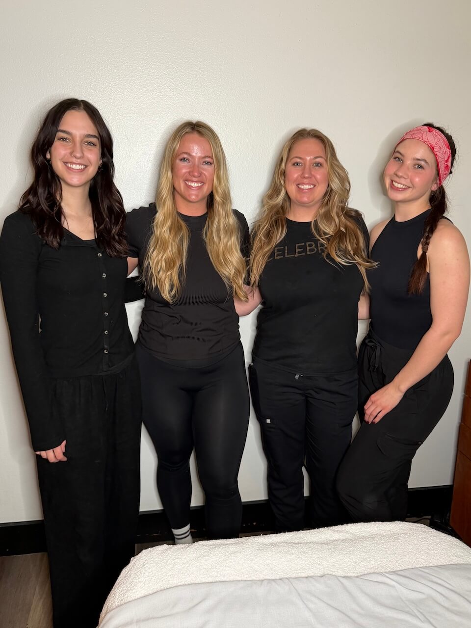 Four women stand side by side smiling in front of a plain light-colored wall, wearing mostly black outfits. Copperfalls Aveda Spa & Salon, Castle Rock, CO