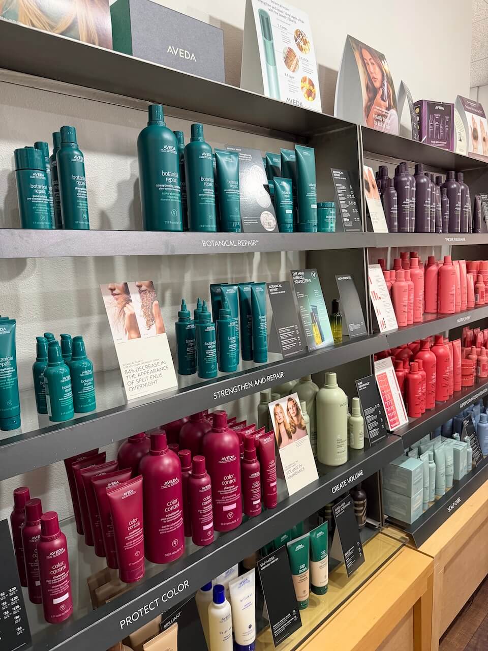 Shelves display Aveda hair care products in various colors and types, organized by function such as protection, repair, and color care, with promotional cards and informational signage. Copperfalls Aveda Spa & Salon, Castle Rock, CO
